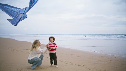 Cheerful kid playing kite with mother on beach. Caring young parent helping son