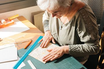 Focused elderly woman sitting at table and applying paper cover on wood hardcover at home