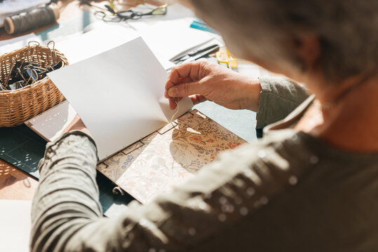 Crop aged woman sitting at table and using needle on notebook in daylight