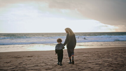 Mother son walking beach holding hands rear view. Carefree family resting sunset