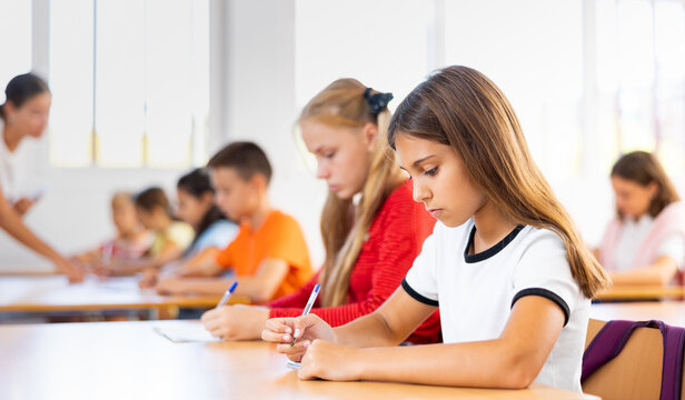 Portrait of interested diligent preteen schoolgirl sitting at school desk and writing exercises at lessons with classmates