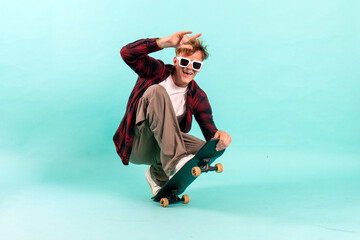 young cheerful guy rides skateboard on blue isolated background, hipster skater in sunglasses shows...