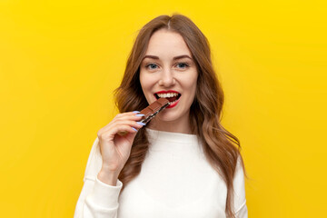 young girl eats and bites big chocolate bar and smiles and rejoices on yellow isolated background