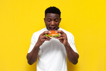 african american man in white t-shirt holding big burger and biting it, the guy eats fast food with his mouth open