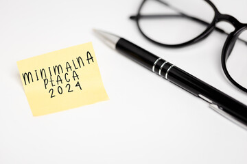 a yellow piece of paper stuck to a white table with the inscription "Minimalna Płaca 2024", next to it are glasses and a pen (selective focus), translation: Minimum Wage 2024