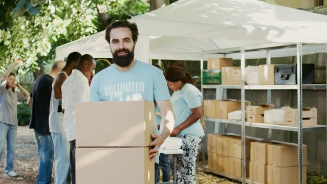 At a food drive, kind young guy holding non-perishable food products for the hungry and homeless. While glancing at the camera, male Caucasian volunteer carries donation packages. Handheld shot.