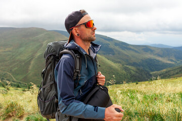 young man in orange jacket and glasses stands on the top of mountain with backpack and trekking...
