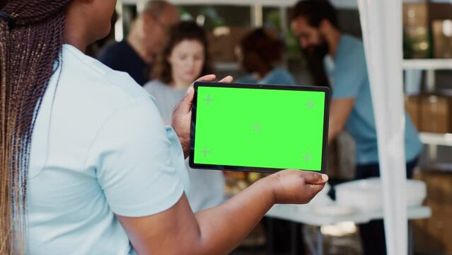 Young black woman clutching digital tablet with isolated mockup template for charitable messaging. African american volunteer carrying smart gadget with blank green screen. Over the shoulder tripod.