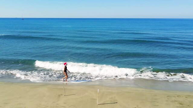 Young beautiful slender woman in Santa Claus hat walking on sandy beach, rejoicing in Christmas or New Year. Shooting with drone. Weekend Sea or Ocean. Resort Vacation.