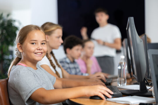 Smiling preteen girl sitting at computer table together with other attendees of IT courses