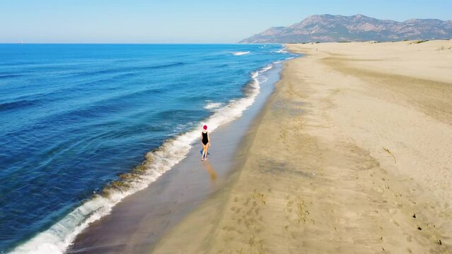 Young beautiful slender woman in Santa Claus hat walking on sandy beach, rejoicing in Christmas or New Year. Shooting with drone. Weekend Sea or Ocean. Resort Vacation.