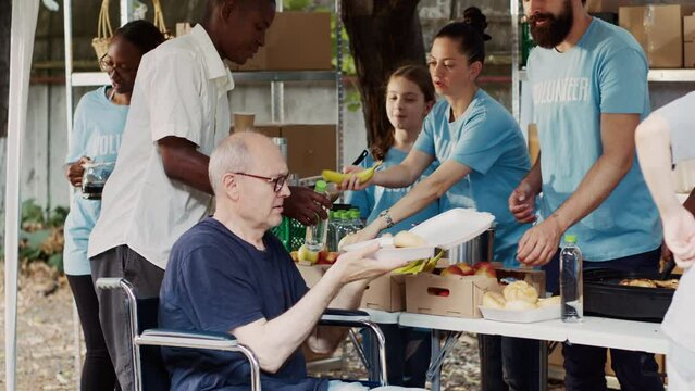 Senior old man in wheelchair receiving free food from group of multiethnic volunteers at hunger relief event. Friendly charity workers providing warm meals to the homeless and disabled people.