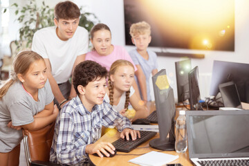 Teenage boy sitting at table and using computer during computer science lesson. Teacher and classmates standing and looking at monitor