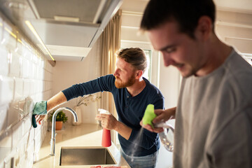 Young male gay couple cleaning their kitchen together - obrazy, fototapety, plakaty