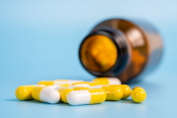 White and yellow pills scattered near opened bottle on blue background. Concept of medical treatment with medicine, vitamins or supplements in capsules, drug addiction