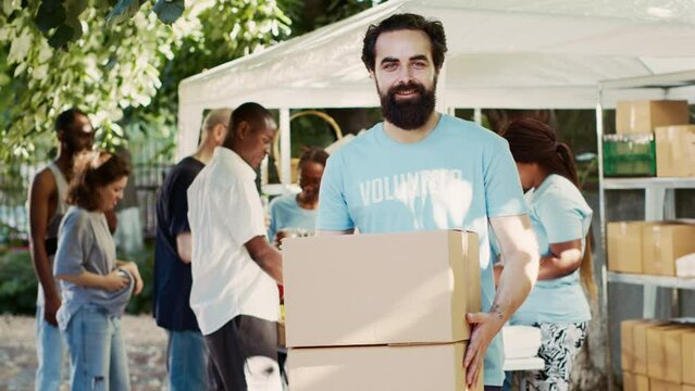 Generous man volunteering at neighborhood food drive, distributing non-perishable rations to the hungry and homeless. While holding donation box, caucasian charity worker looks at camera. Tripod shot.
