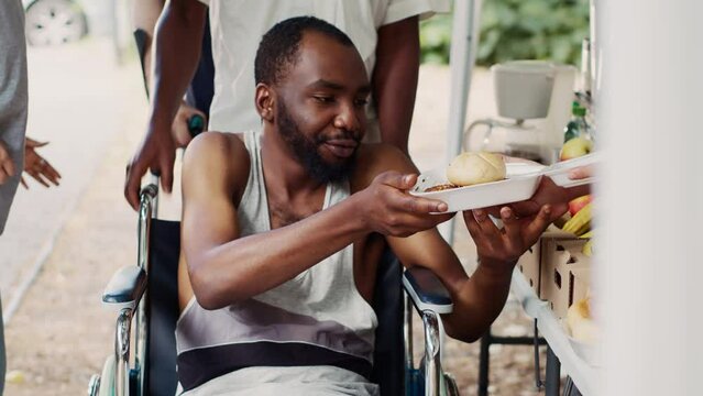 Free meals and fruits are given to a wheelchair-bound african american man in need. Handicapped, poor and homeless people receive provisions from a non-profit food drive. Close-up, tripod shot.