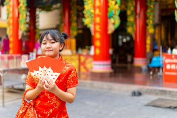 Portrait of Happy Little Asian girl in Chinese red dress holding red envelopes in front of in Chinese temple shrine. Chinese lunar new year festival, holiday celebration, religion meditation concept.