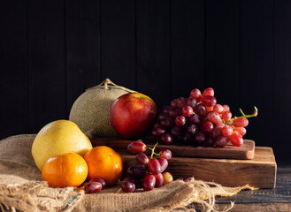 Orange fruits, grapes, apples, and cantaloupe are placed on a wooden table, black and dark background.