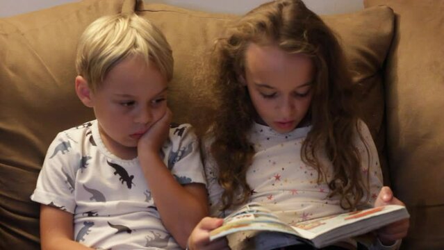 LITTLE SISTER READS TO LITTLE BROTHER WHILE HE PICKS HIS NOSE