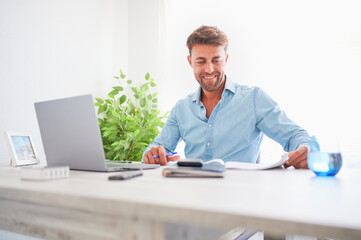 Smiling young confident man dressed in blue working with a computer at his workplace in the office. Happiness at work concept.