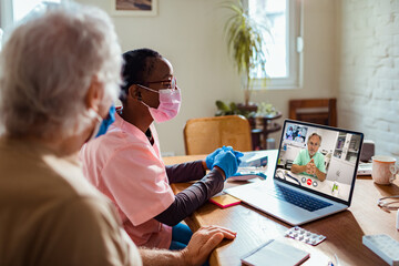 Young African American caregiver consulting a doctor on a video call on the laptop with her senior patient at home