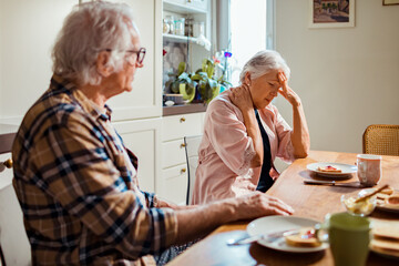 Senior Caucasian woman is having neck pain while eating breakfast with her husband in the morning in the kitchen