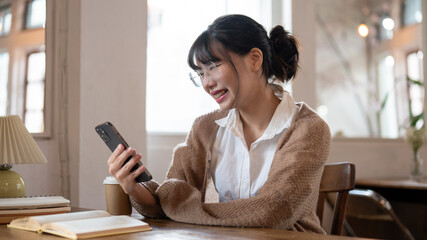 A happy Asian woman reading messages on her smartphone while sitting at a table indoors.