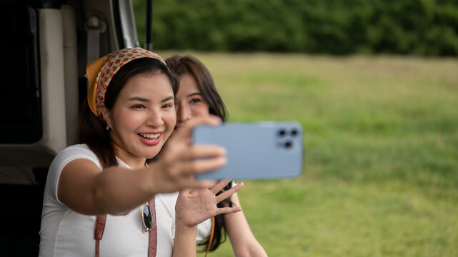 Two pretty Asian female friends are sitting in the car's open trunk, using a phone to take selfies