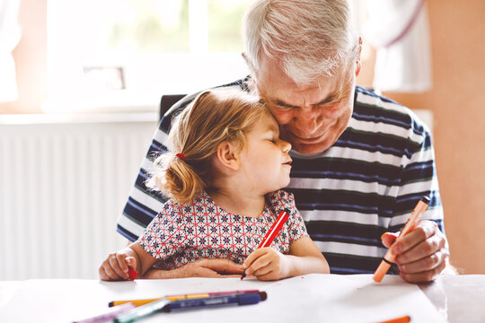 Cute little baby toddler girl and handsome senior grandfather painting with colorful pencils at home. Grandchild and man having fun together