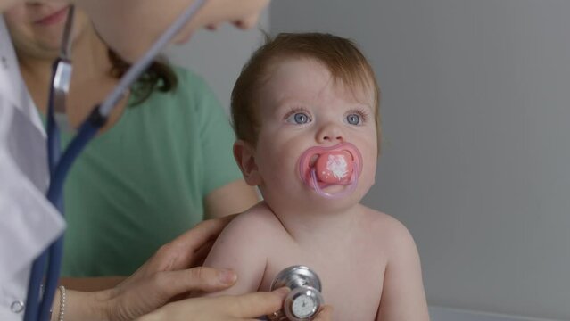 Cute Baby Sits On Changing Table, Takes Out Pacifier From Mouth, Waves Hands, Cries. Female Pediatrician Tries To Listen Heartbeat Of Kid Using Stethoscope. Mom With Little Child On Checkup. Close Up.