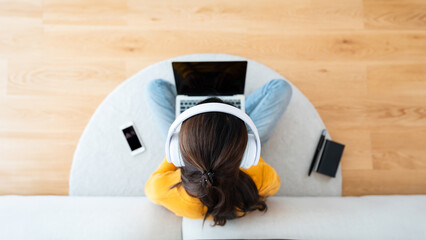 Top view of young Asian woman sitting on the floor and working on computer laptop with book, pen and mobile smartphone, wearing wireless headset, listening to music - Powered by Adobe