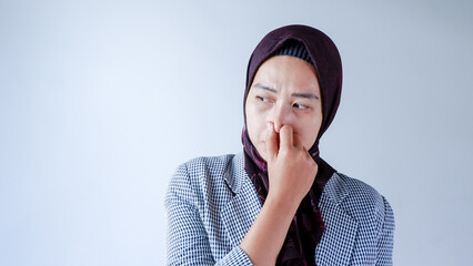 Portrait of frustrated Asian woman wearing hijab and formal wear pinching nose with disgust on his face due to bad smell isolated over gray background. - obrazy, fototapety, plakaty