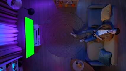 Top view of male resting on the couch in the room playing video game with joystick, TV with Chroma key green screen, advertising area workspace mockup.