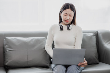 Asian woman surfing social media with headphones using laptop at home on sofa