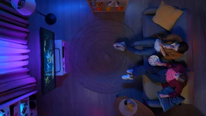 Fotobehang Gaming Top view of couple sitting on the couch in the living room, watching movie on TV eating popcorn.  © kinomaster