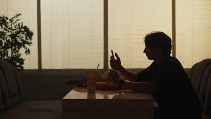 Silhouette of young woman sitting at the bar, holding smartphone watching social media eating pizza.