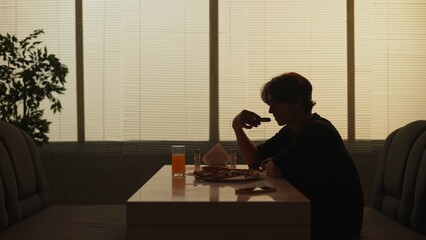 Silhouette of young man sitting at the bar, male on a lunch time break alone, eating holding pizza slice.