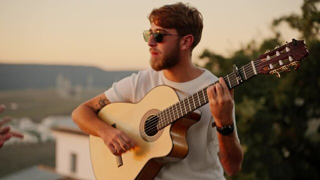 Solo energetic young man playing electric guitar with hand fingers while wearing sunglasses. Adult boy performing guitar excited, confident. View from top of building, sea beach water in beckground
