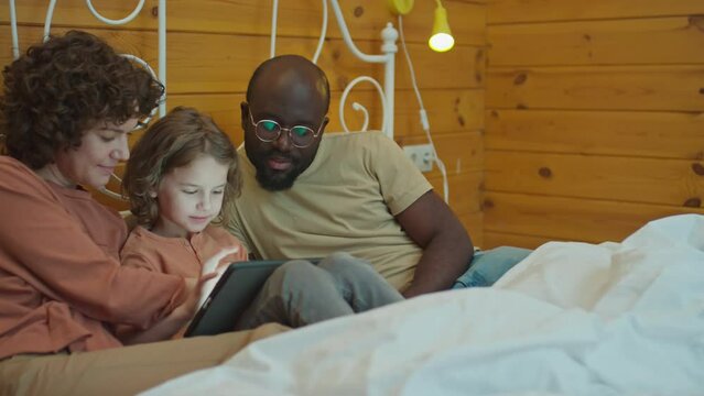 Medium shot of interracial parents and their child in bedroom sitting on bed looking at photos on tablet