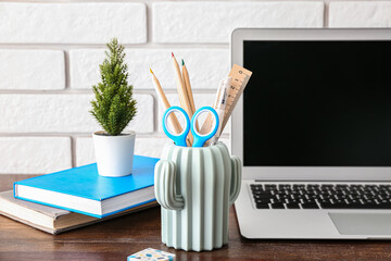 Modern laptop, houseplant and holder with different stationery on wooden desk, closeup