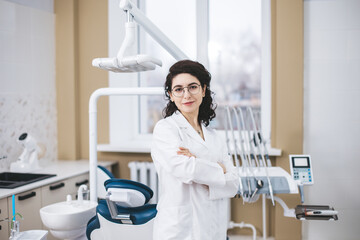 Fototapeta premium Professional young dentist posing confidently beside modern dental equipment in a dental clinic. Portrait of female doctor.