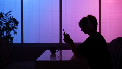 Silhouette of man sitting at the food bistro bar at night in neon light, wearing headphones, listening music.