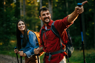 A multicultural pair with backpacks and walking sticks discover the beauty of the outdoors, their beaming smiles mirroring their excitement for the journey ahead - obrazy, fototapety, plakaty