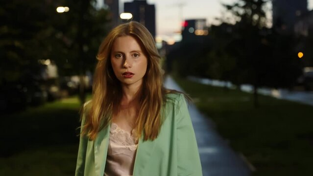 Fashionable gait of a beautiful girl in a night summer park. A girl with long hair has a green jacket and a beautiful look. Night cityscape. A dynamic shot.
