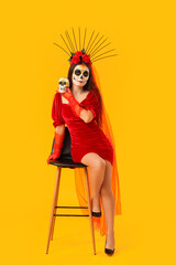 Young woman with painted skull sitting on yellow background. Mexico's Day of the Dead (El Dia de Muertos) celebration