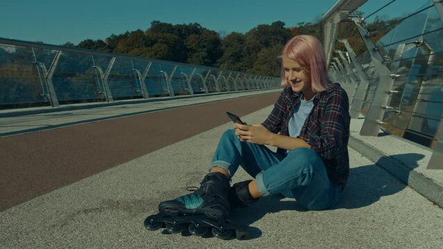 Positive pretty pink haired woman in roller skates sitting on pedestrian bridge, networking and browsing social media content online using cellphone while rollerblading in city street at daybreak.