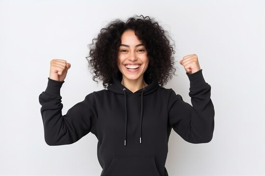 Cheerful young african american woman in black hoodie celebrating success, clenching fists and smiling. Isolated on white background
