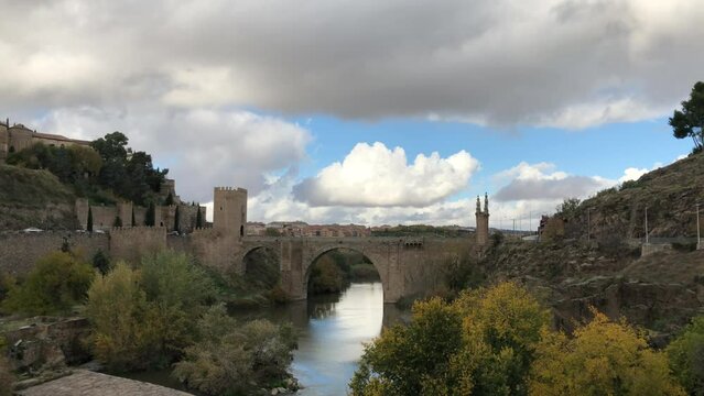 Shot of Alcantara Bridge over Tagus river, reflecting white cotton clouds and blue sky. Roman & medieval style arch bridge, Toledo, Spain. Slow atmosphere.