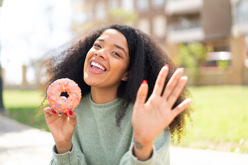 Young African American woman holding a donut at outdoors saluting with hand with happy expression